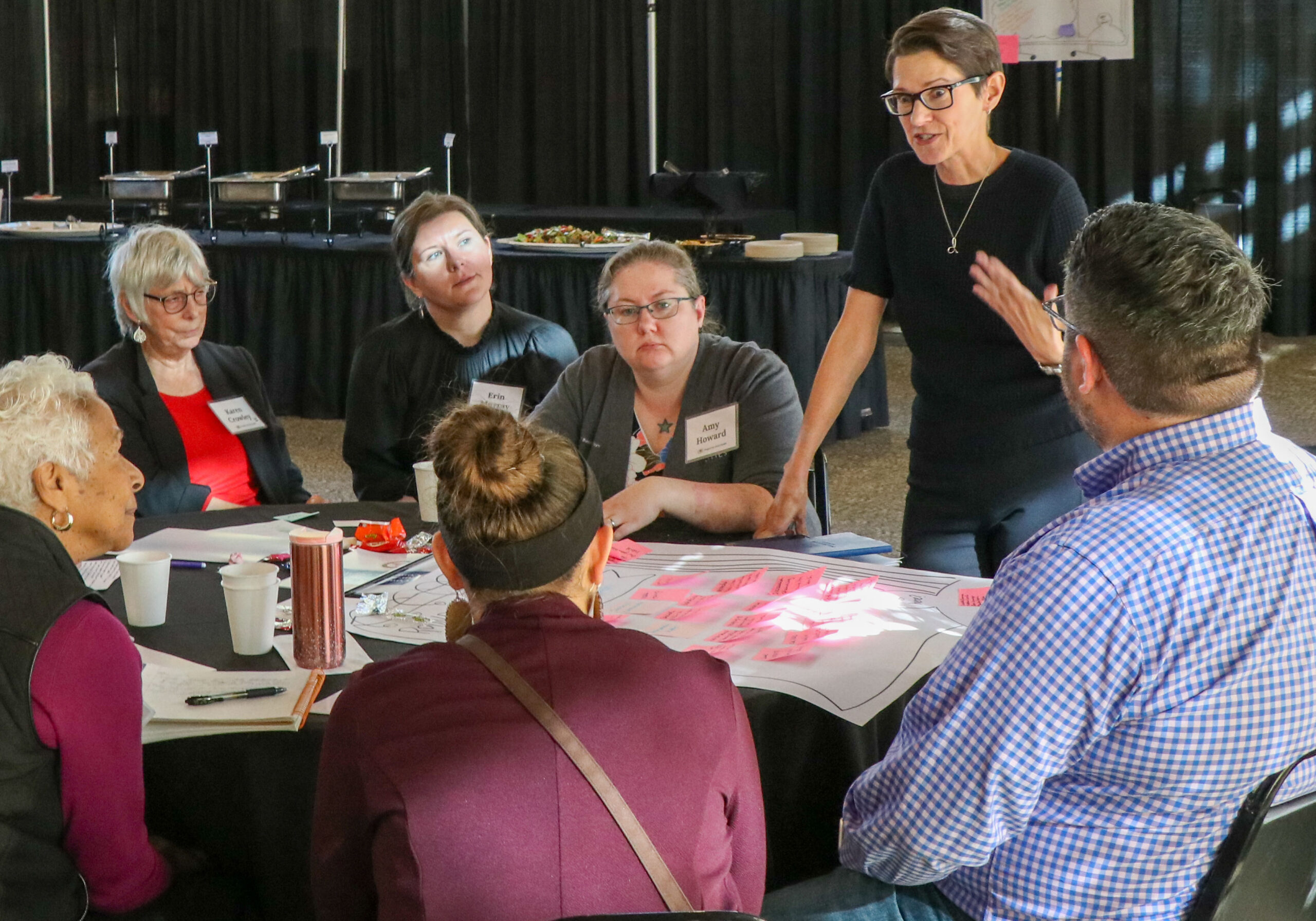 A woman talks to a group of people seated at a table while standing above an easel-sized sheet of paper with stickie notes on it