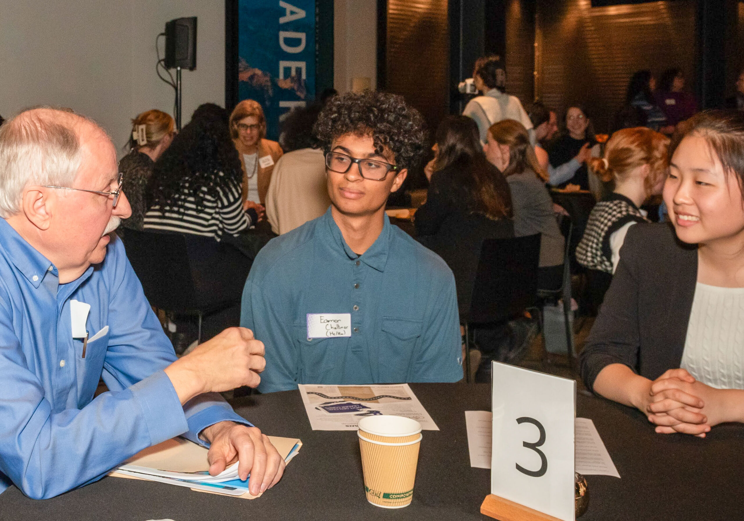 A man talks to two students who are seated at a table with him
