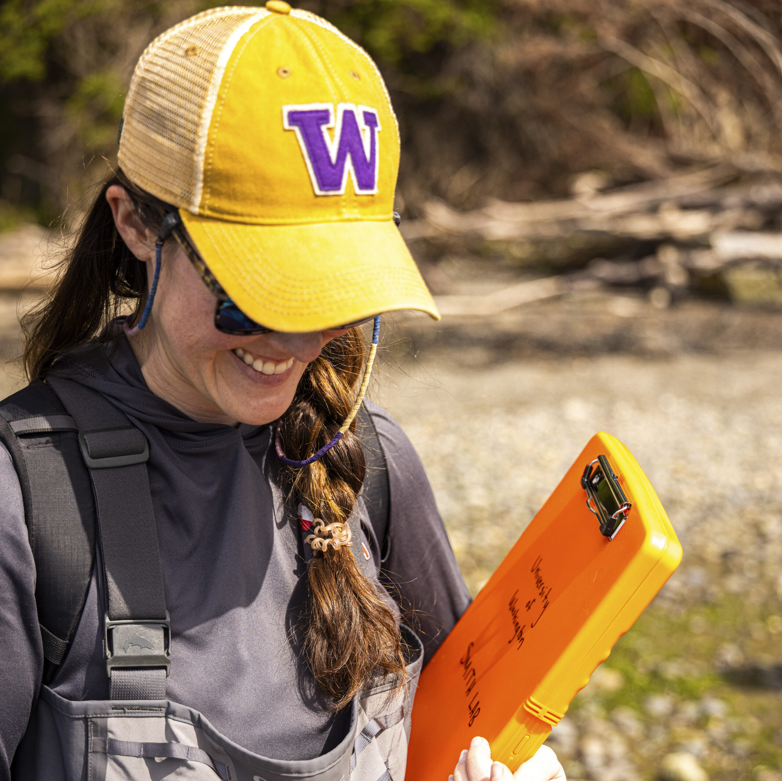 A student wearing a gold University of Washington hat holds an orange clipboard while surveying a tide flat